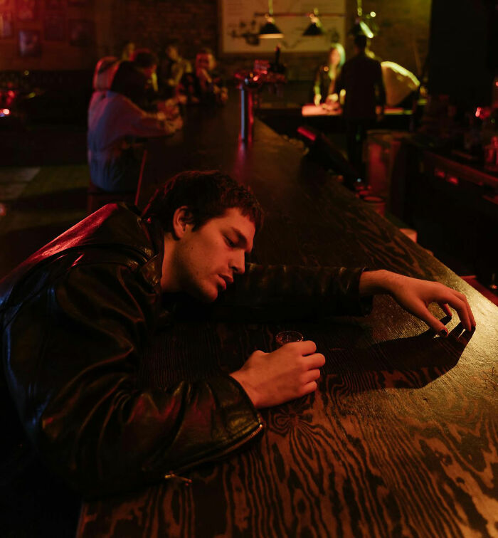 Man sleeping at a casino bar, holding a shot glass, with dim lighting and other patrons in the background.