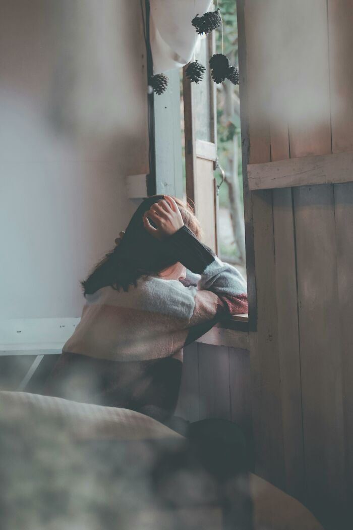 Person in a cozy sweater leaning on a windowsill, gazing outside, reflecting on secrets kept from loved ones.