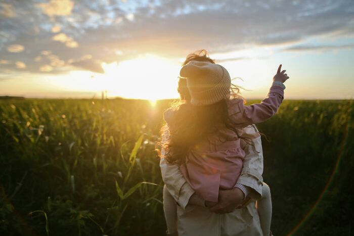 Person holding a child in a field at sunset, both facing the horizon.