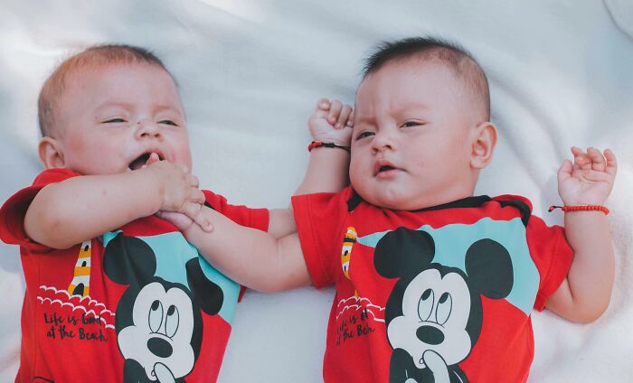 Two babies in matching red Mickey Mouse shirts lying on a white blanket, sharing a playful moment.