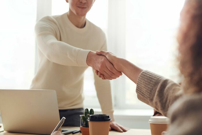 Two people shaking hands in a peaceful setting, with coffee cups and a laptop on the table.