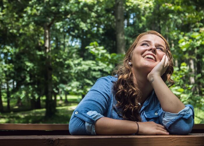 Woman in a blue shirt smiling thoughtfully, captures moment realized dating idiot, in a lush green forest background.