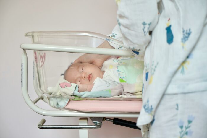 Baby sleeping in a hospital crib with a person's hand gently touching its head.