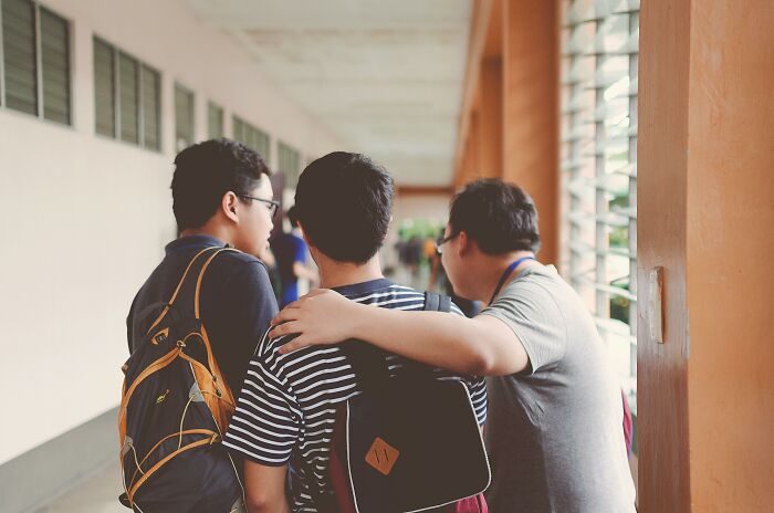 Three friends in a hallway, one with an arm around another, discussing unexpected "You Up?" text outcomes.