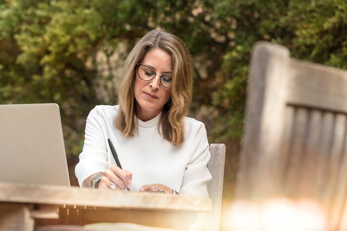 Woman in glasses writing at an outdoor desk with a laptop, representing the concept of getting fired quickly.