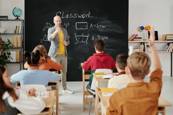 Teacher in a classroom instructing students, with geometric formulas on the blackboard, discussing worst jobs ever had.