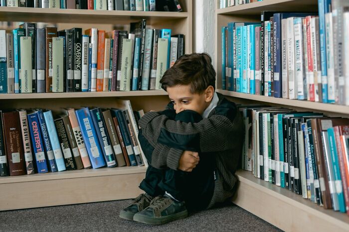 Child sitting alone in a library corner, surrounded by bookshelves, looking contemplative.