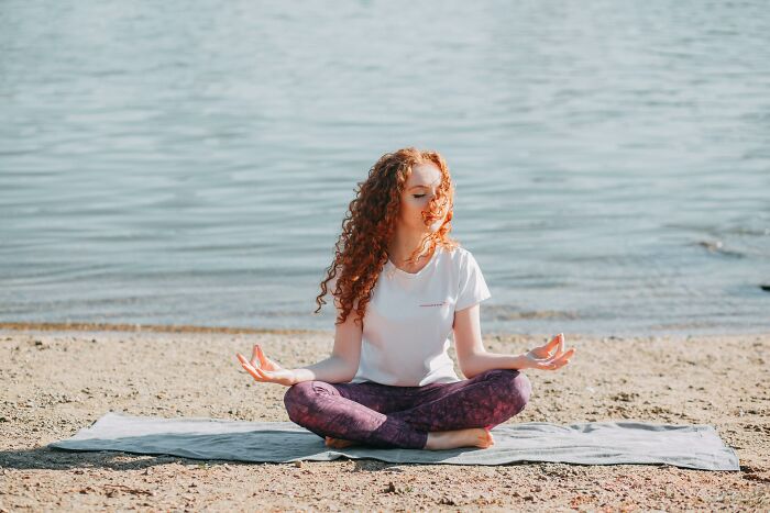 Person meditating on a beach, wearing a white shirt and purple leggings, focusing on relaxation and mindfulness.