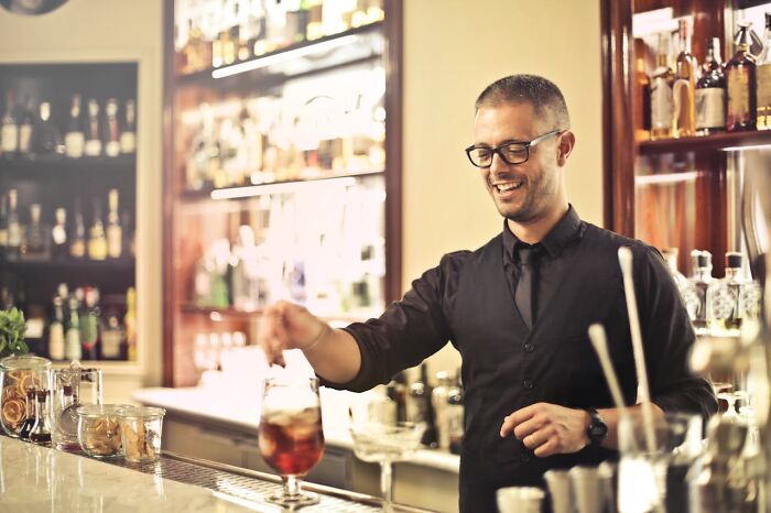 Bartender in a black vest serving a drink at a bar, smiling, with various bottles in the background.