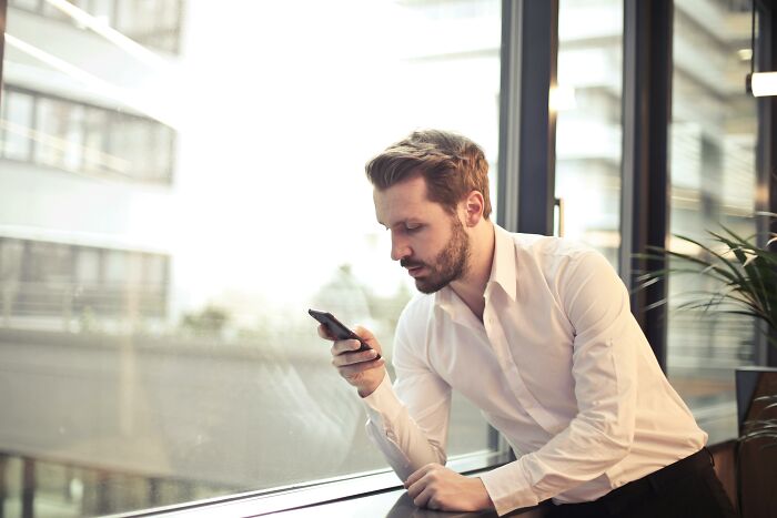 Man in office attire looking at smartphone by a large window, related to the "WomenInMaleFields" trend.