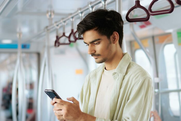 Man in a light green shirt on a train, looking at his phone, experiencing an astonishing coincidence.