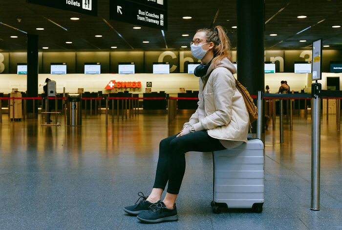 Traveler wearing a mask, sitting on a suitcase at an airport terminal, facing a closed check-in area.