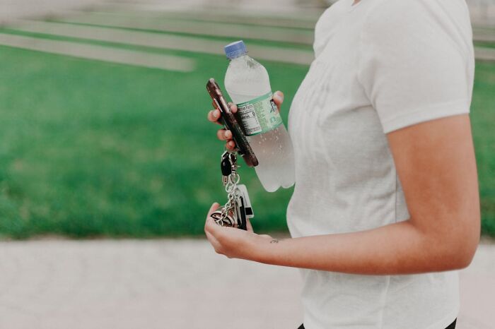 Person in a white shirt holding a water bottle, phone, and keys, emphasizing hygiene habits outdoors.