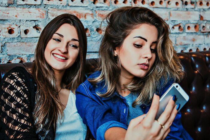 Two women sitting on a couch, one taking a selfie and the other smiling, highlighting social interactions for secret judgement.