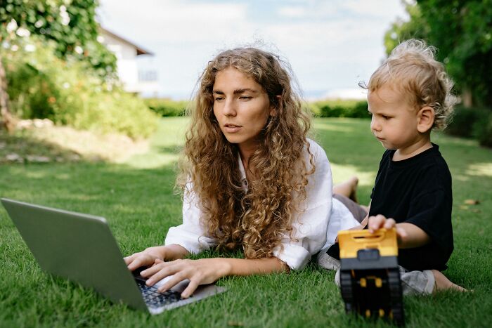 Woman enjoying peace and quiet while working on a laptop outdoors with a child playing nearby.