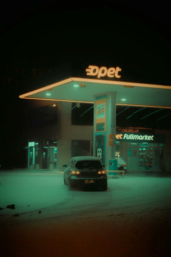 Car parked at a snowy gas station at night, illuminated by green and orange lights, depicting a quiet and isolated scene.