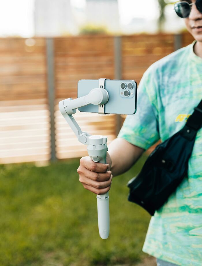 Person holding a smartphone on a gimbal in a casual outdoor setting, wearing sunglasses and a tie-dye shirt.