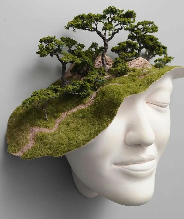 Sculptor's face sculpture blending realism with surrealism; features trees and paths on head.