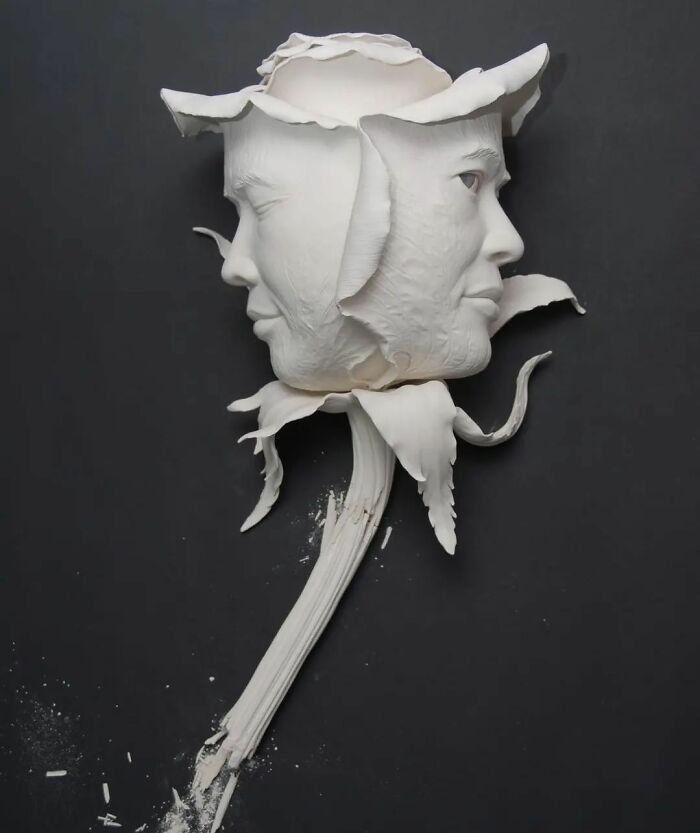 Surrealism and realism blend in a sculpted white rose with human faces against a black background.