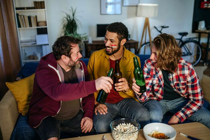 Three friends sitting on a couch, smiling and clinking beer bottles in a cozy living room setting.