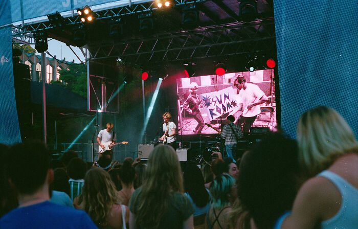 A band performing live on stage at an outdoor concert with a screen showing the band and a crowd watching.