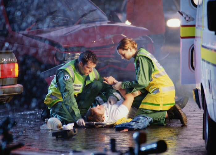 Paramedics assisting an injured person after a car accident at night, with emergency lights flashing and rain falling.