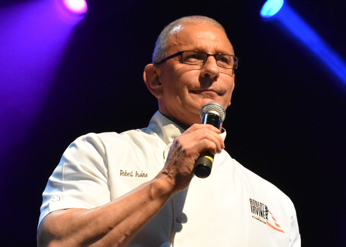 Chef speaking into a microphone on stage, wearing a white chef's jacket, under stage lights.