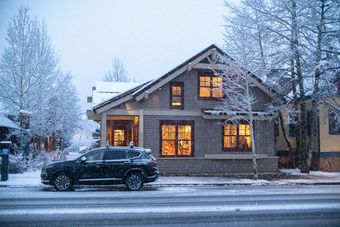 Snow-covered house with warm interior lights, trees, and parked car, reflecting winter scene in a neighborhood.