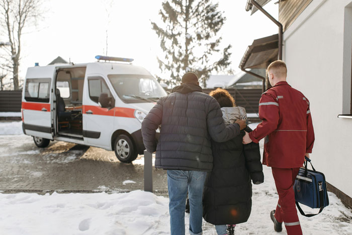 Paramedic assists two people walking through snow towards an ambulance.