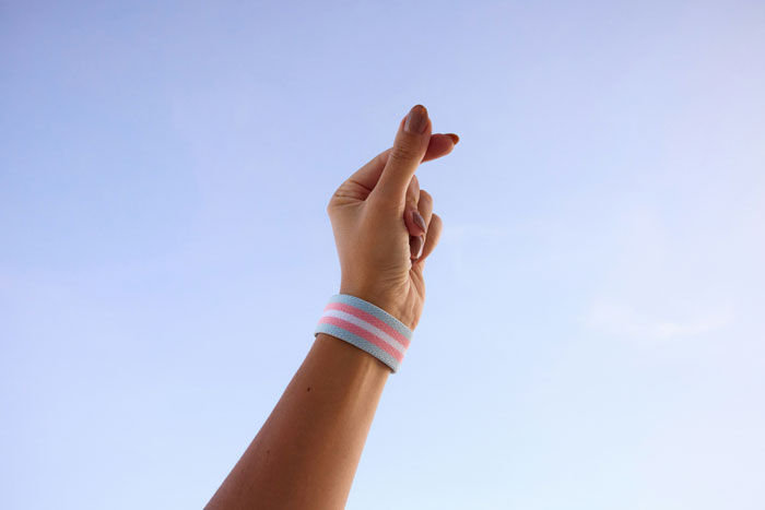 A hand with a pink and blue wristband raised against a clear sky, symbolizing transgender pride and identity.