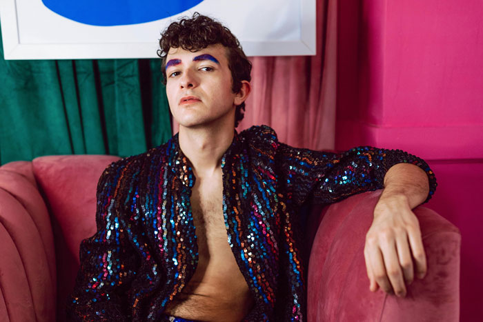 Person with blue eyebrows in a colorful sequin jacket, sitting on a pink couch.