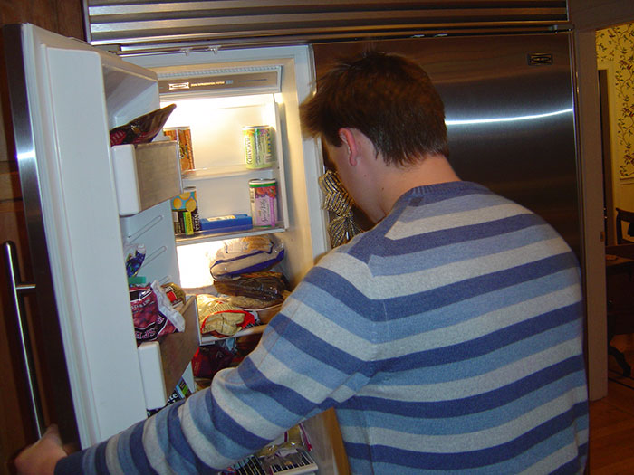 Man in a striped sweater looking into a refrigerator, referencing awkward living situation with his girlfriend.