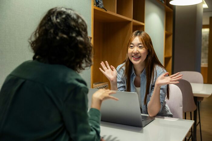 Two women chatting at a table with a laptop, possibly discussing things people secretly judge others for.