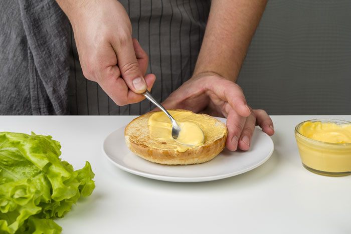 Spreading mayonnaise on a bun; examining its consistency with nearby lettuce and bowl of mayo.