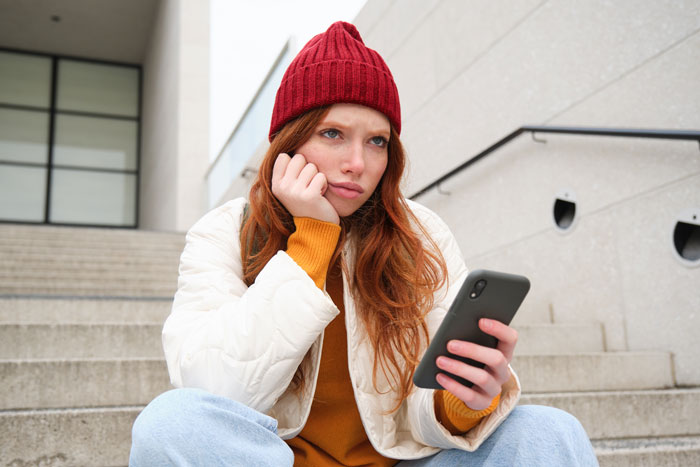 Woman sitting on stairs, wearing a red beanie, looking concerned while holding a phone.