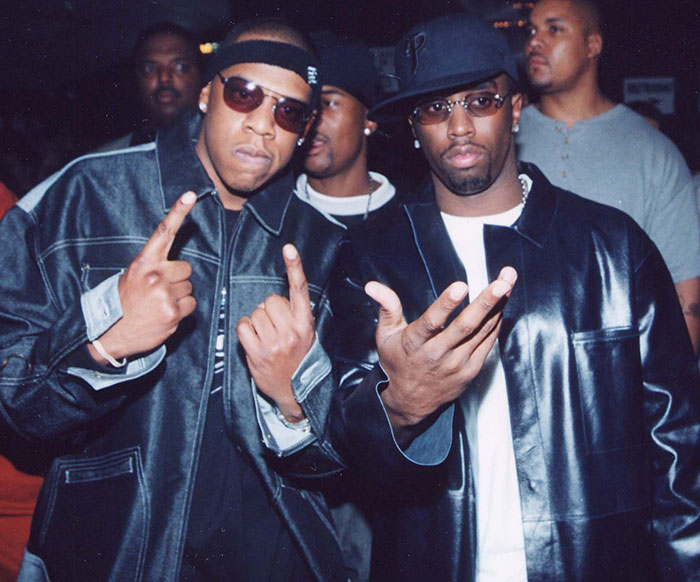 Rappers Jay-Z and P Diddy in sunglasses and leather jacket reacts at a public event, gesturing with his hand.