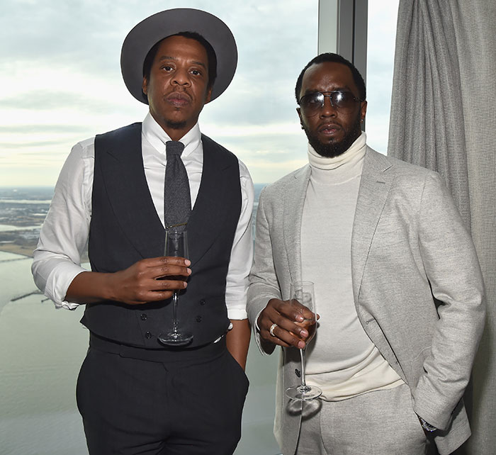 Jay Z and Sean Diddy Combs in formal attire stand by a window with drinks, involved in a legal controversy.