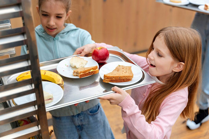 Two young girls holding lunch trays, selecting food items in a cafeteria.