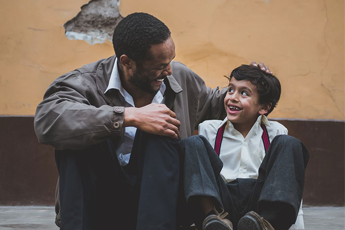 A smiling father and son sit against a wall, depicting positive parenting dynamics.