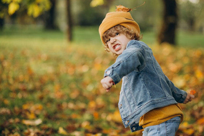 Child in a denim jacket and orange hat showing frustration in an autumn park.