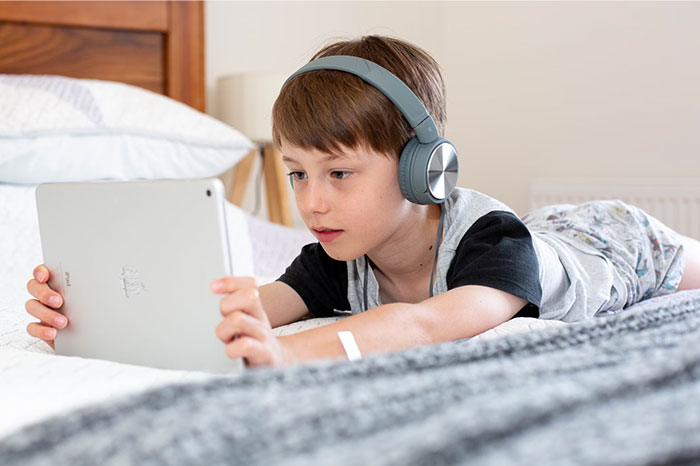 Child wearing headphones, focused on a tablet, lying on bed, illustrating a sign of terrible parents.