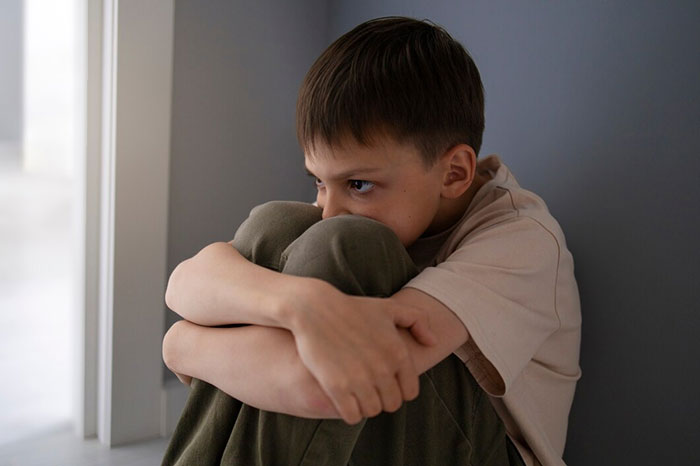 A boy sitting alone in a corner, hugging his knees, exhibiting signs of terrible parents.