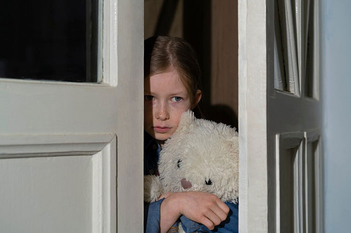 Child holding a teddy bear, looking worried behind a partially open door; signs of terrible parents concept.