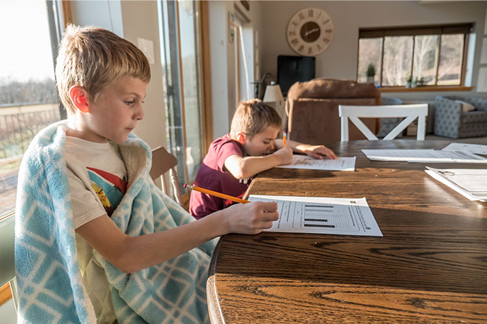 Two children doing homework at a table, one wrapped in a blanket, in a sunny home setting, illustrating signs of terrible parents.