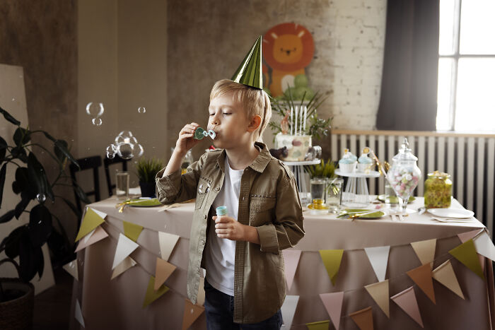 Boy wearing a party hat blows bubbles near a decorated table during a festive event.