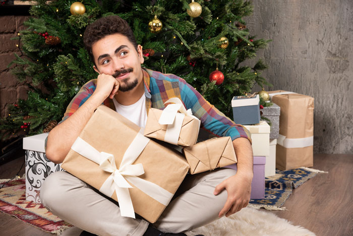 Person sitting with gifts by a Christmas tree, contemplating debt clearance for 1642 people using $5k intended for family gifts.