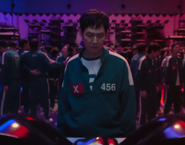 A contestant in a green tracksuit from Squid Game 2 stands in a dimly lit room with others.