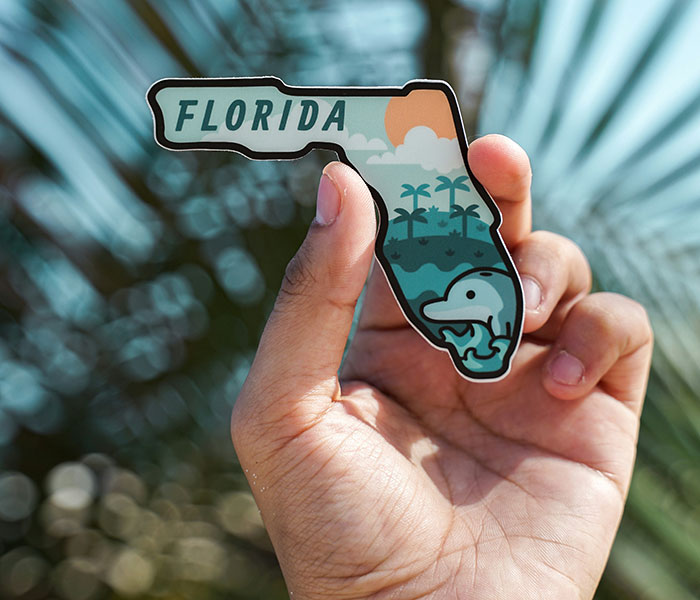 Hand holding a Florida-shaped sticker, featuring palm trees, the sun, and a dolphin design.