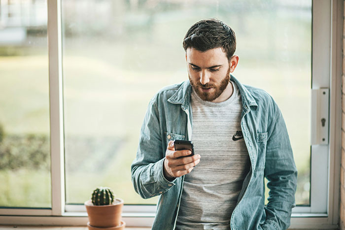 Man in a denim shirt looking at his phone by a window, embodying a casual Canadian style.