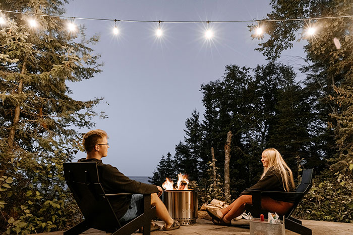 Two people sitting by a fire pit under string lights, surrounded by trees, enjoying a Canadian evening outdoors.